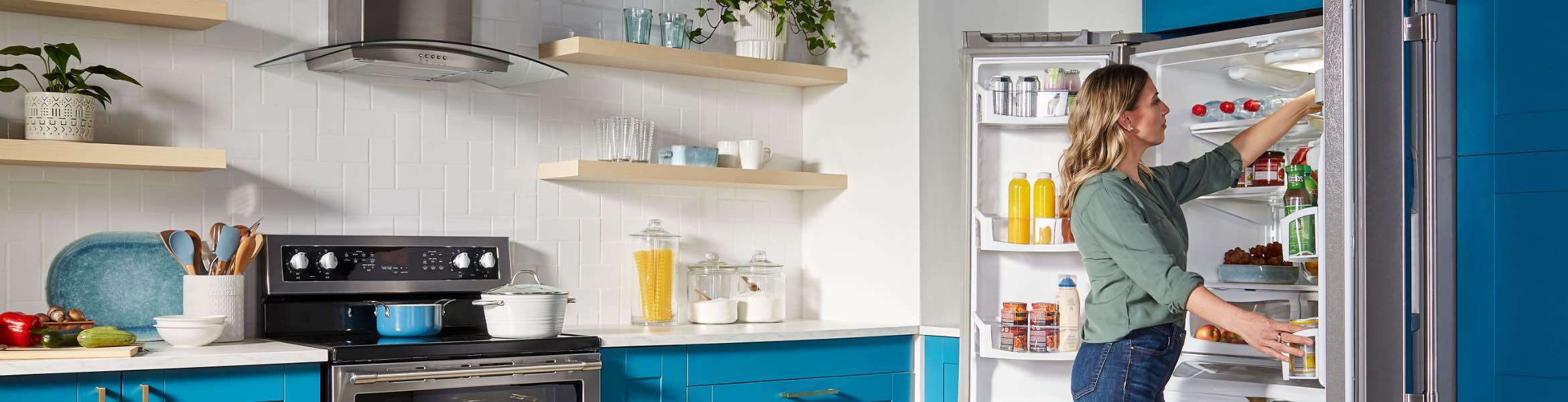 A woman in front of an opened refrigerator. Inside the fridge are beverage bottles, condiments, bottles of dressing, water bottles and more, and she is surrounded by blue drawers and cabinets.