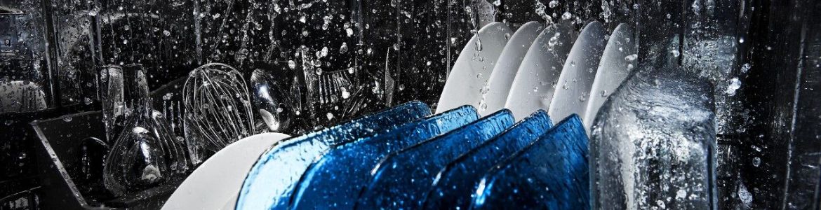 Inside a Maytag dishwasher during a wash cycle. A baking dish, plates and a cutlery tray filled with cutlery and utensils are being cleaned.