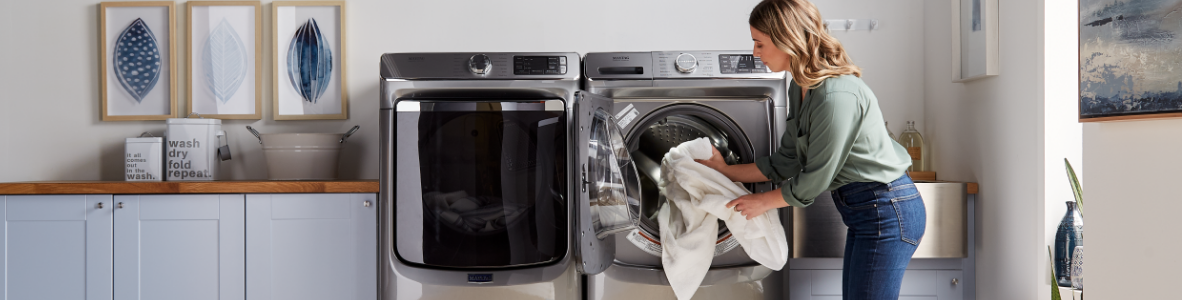 A woman in jeans and a green blouse loads linens into a Maytag Dryer. Next to the appliance is a Maytag Washer and next to the woman is a basket of laundry.