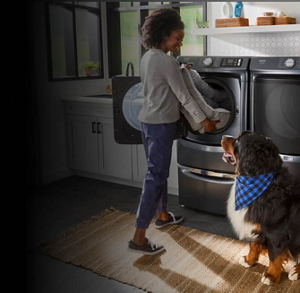 A dog watching its owner loading a dog bed into a front load washer on a pedestal next to a front load dryer