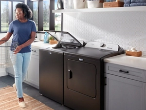 A woman carries a laundry basket to her Maytag washer and dryer, which are surrounded by cabinets with marble tops. The lid on the washer is open.